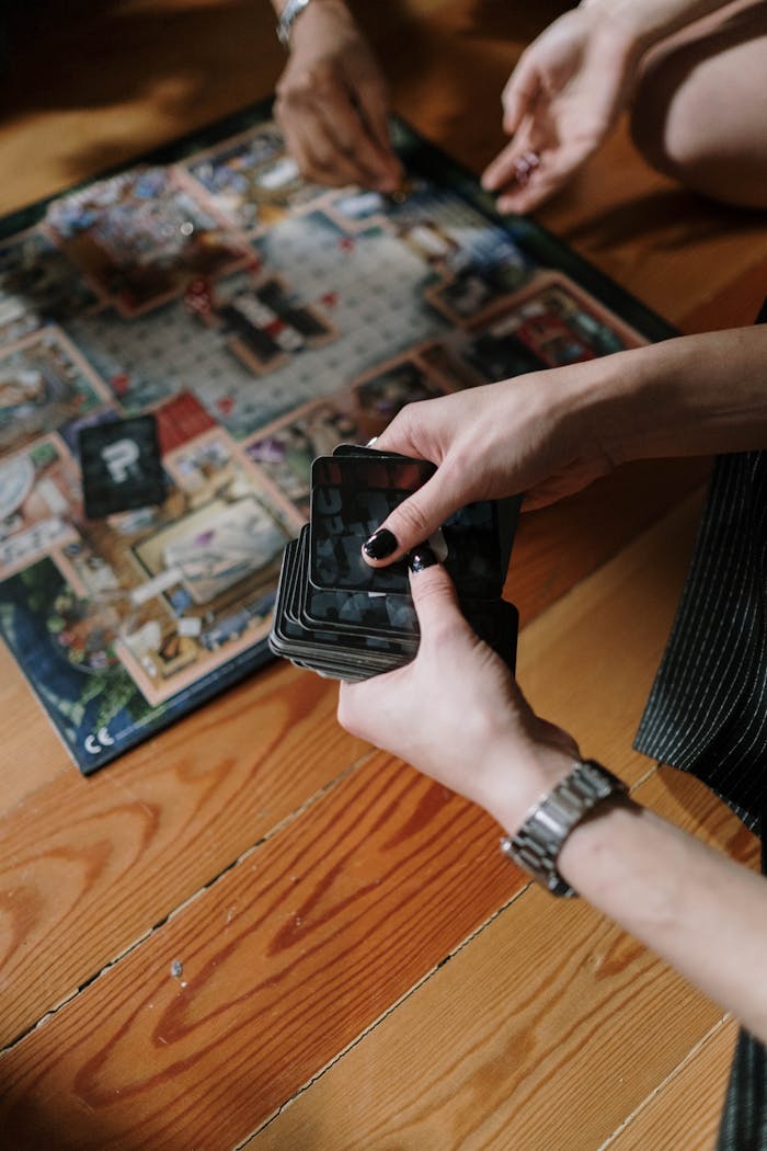 Hands holding cards during a board game session on a wooden floor indoors.